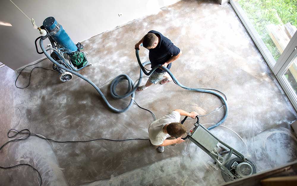 Two men working on a floor renovation, one operates a grinding machine while the other manages the vacuum hose. Light fills the room.
