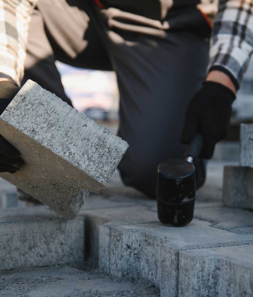 A person in gloves holds a concrete block above a paved area, preparing to place it while a mallet rests nearby.