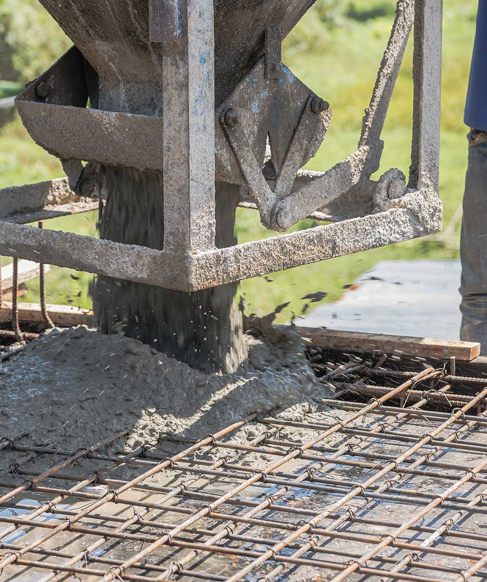 A concrete mixer pouring fresh cement onto a rebar grid, with a worker’s leg visible, set against a green landscape.