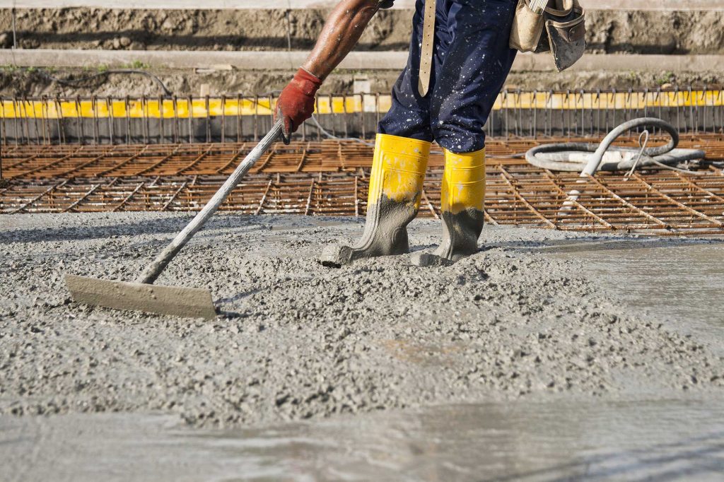 A worker levels wet concrete on a construction site, wearing gloves and rubber boots. The background shows reinforcing steel bars and other construction materials.