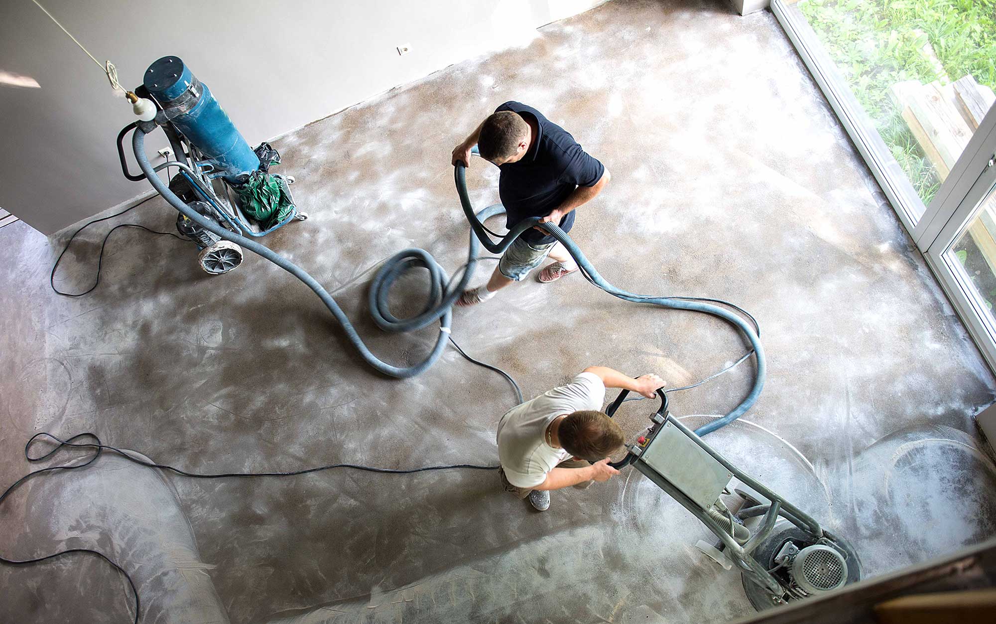 Two workers are using industrial floor cleaning equipment on a concrete surface. The scene is viewed from above, showing the tools and the process of floor maintenance.