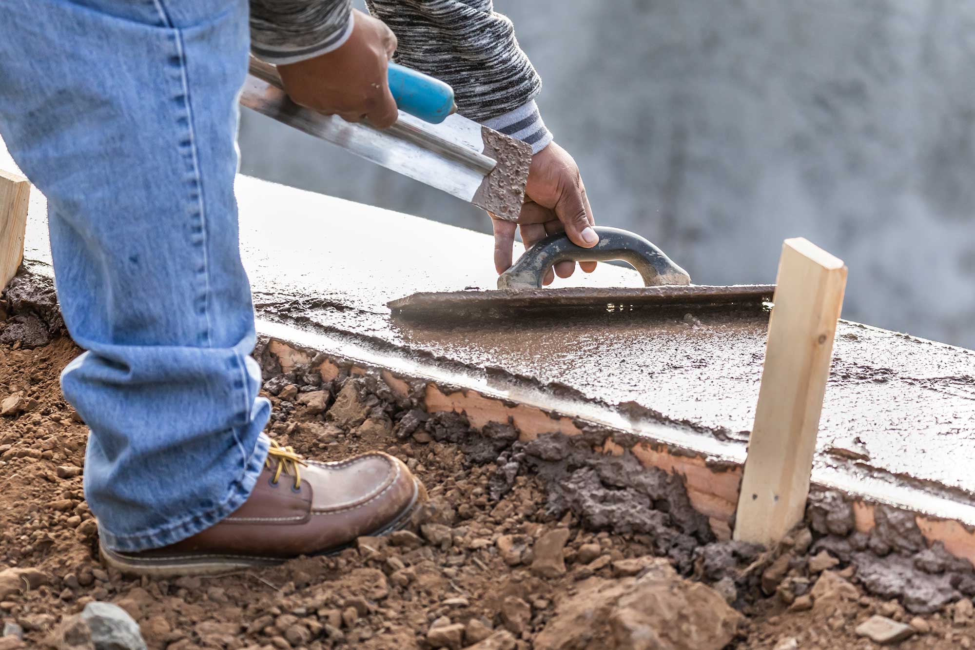 A worker smooths wet concrete with a trowel on a construction site, surrounded by wooden forms and soil.