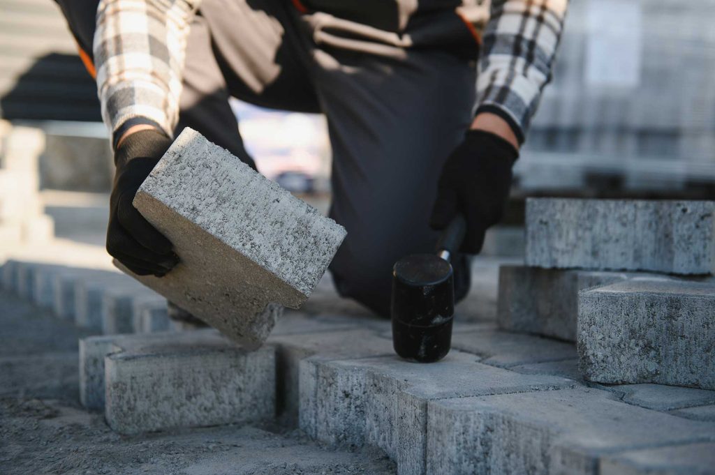 A person wearing gloves is lifting a stone block while working on a construction site, surrounded by stacked stones.