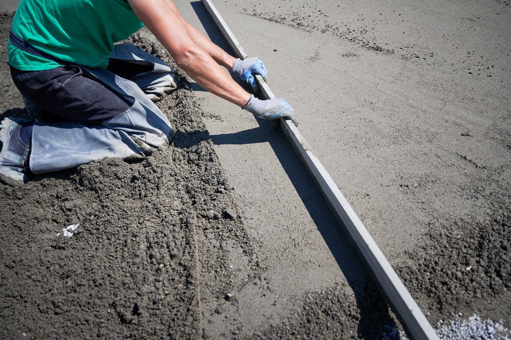 A worker levels freshly poured concrete using a straightedge tool. The scene captures the process of concrete finishing.
