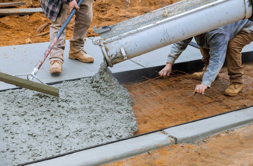 Construction workers are pouring concrete onto a prepared surface, using tools to spread it evenly. The scene captures the process of laying a concrete slab.