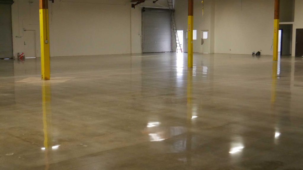 A spacious industrial interior with a polished concrete floor and yellow support columns, featuring a large garage door in the background. The area is well-lit and empty, suggesting it may be used for storage or manufacturing.