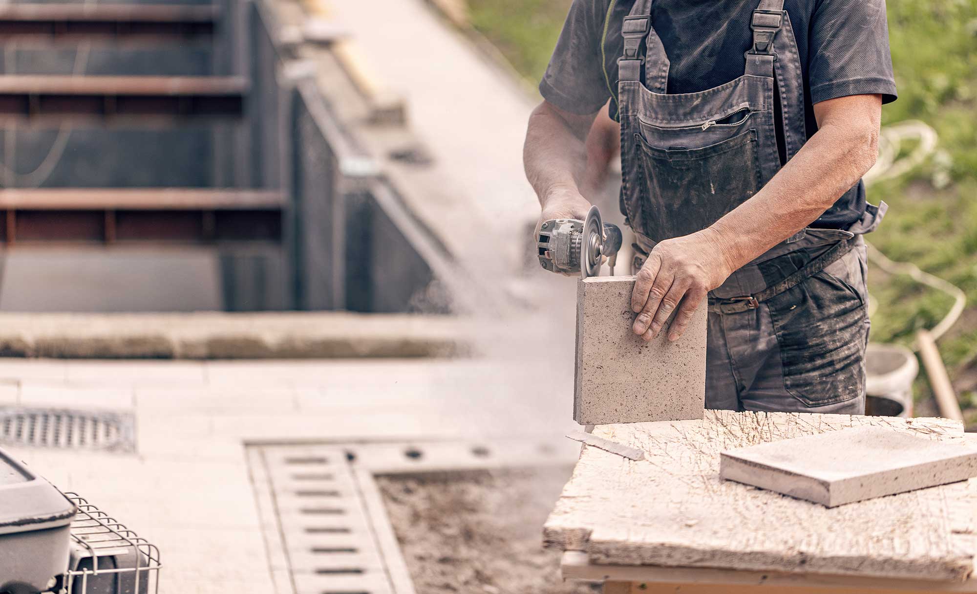 A construction worker uses a power tool to cut a stone slab on a building site. Dust is visible in the air, indicating active work.
