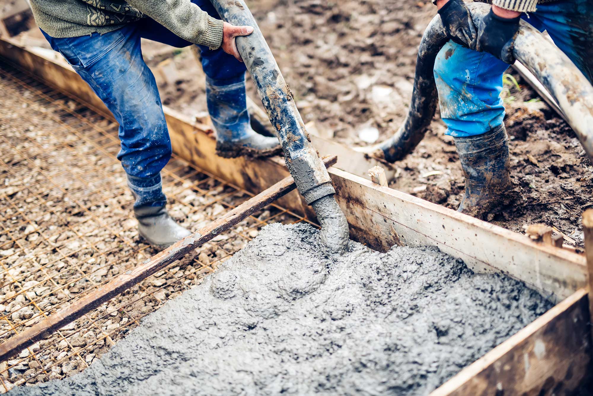 Workers are pouring concrete into a wooden form using a hose. The scene depicts a construction site with muddy surroundings.